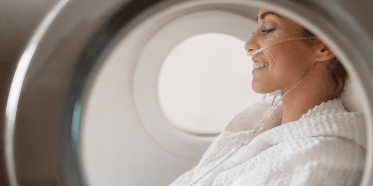 A woman smiles as she sits inside a hyperbaric oxygen chamber, receiving oxygen-rich therapy.
